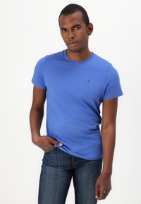 Camiseta de algodón azul con cuello redondo, mangas cortas y pequeño logo en el pecho. Combinada con jeans de mezclilla oscura.