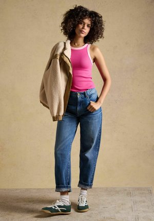 Kvinde med krøllet hår iført en lyserød tanktop, blå jeans, hvide sokker og grønne sneakers, med en beige jakke over den ene skulder.