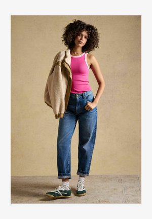 Kvinde med krøllet hår iført en lyserød tanktop, blå jeans, hvide sokker og grønne sneakers, med en beige jakke over den ene skulder.