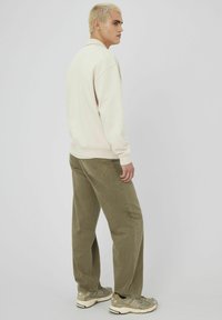 Sweatshirt beige avec des poignets et un ourlet côtelés, associé à un pantalon droit olive. Le modèle porte des baskets grises avec des accents subtils.