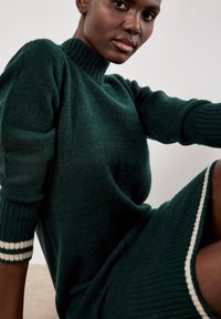Abito lavorato a maglia di un verde scuro con colletto alto a coste, maniche a tre quarti e dettagli a righe bianche sui polsini e sull'orlo.