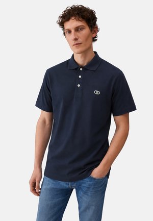 Uomo che indossa una polo blu navy a maniche corte con bottoni bianchi e jeans chiari, in piedi con una mano in tasca.