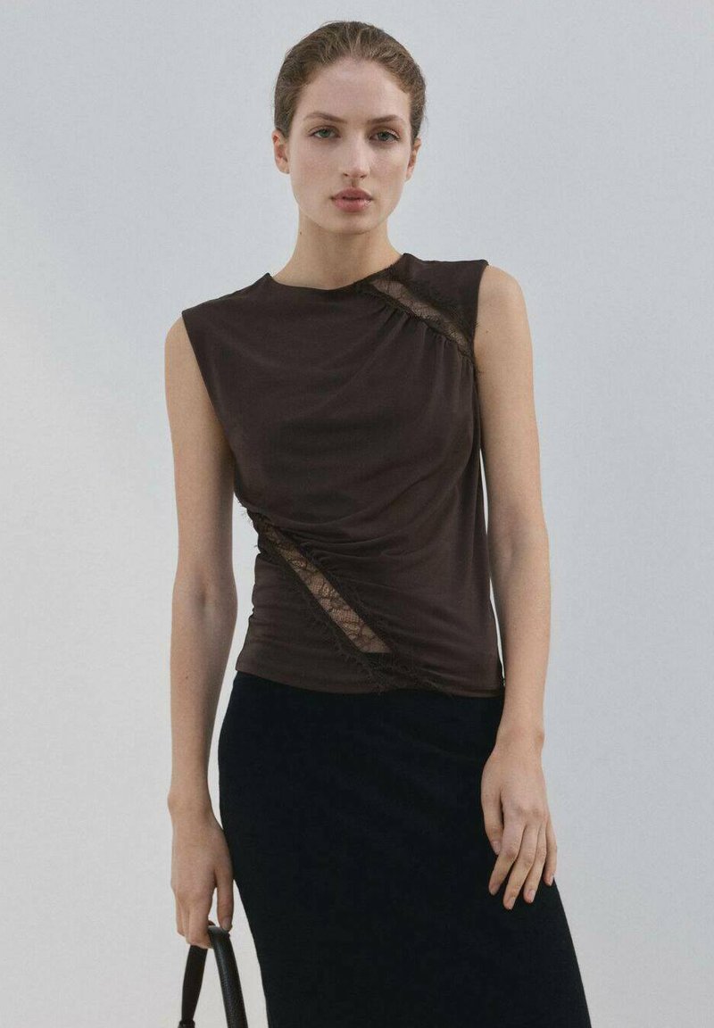 Žena u bezrukom crnom topu s dijagonalnim čipkastim detaljima i crnoj suknji, držeći crnu torbicu na jednobojnoj pozadini.