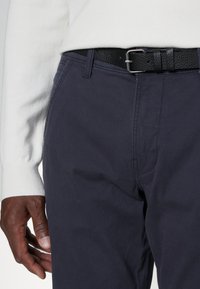 Tamsiai mėlynos medvilninės kelnės su lygia tekstūra, turinčios subtilius siuvinėjimo detalių ir juodą odinį diržą su tekstūruotu paviršiumi.