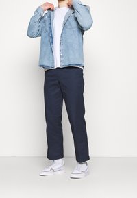 Jasnoniebieska koszula z guzikami na linii klatki piersiowej nałożona na białą koszulkę, w połączeniu z ciemnogranatowymi spodniami i białymi sneakerami. Koszula ma dwie kieszenie na piersi.