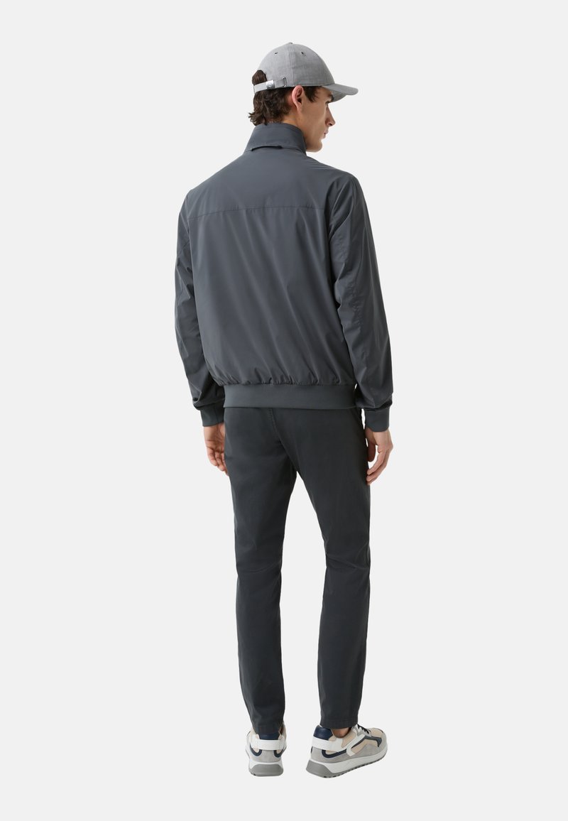 Leichte graue Bomberjacke für Herren mit hohem Kragen, elastischen Bündchen und Saum, kombiniert mit dunklen Hosen und grauen Sneakern.