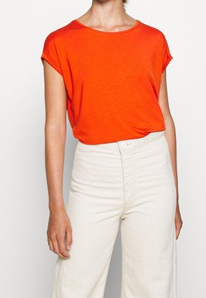 Person trägt ein helloranges kurzärmeliges Hemd, das in hochtaillierte, cremeweiße Hosen gesteckt ist, vor einem einfarbigen weißen Hintergrund.