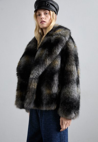 Femme blonde portant un manteau en fourrure multicolore foncé, une casquette en cuir noire et un pantalon bleu marine à motifs, se tenant devant un fond gris uni.