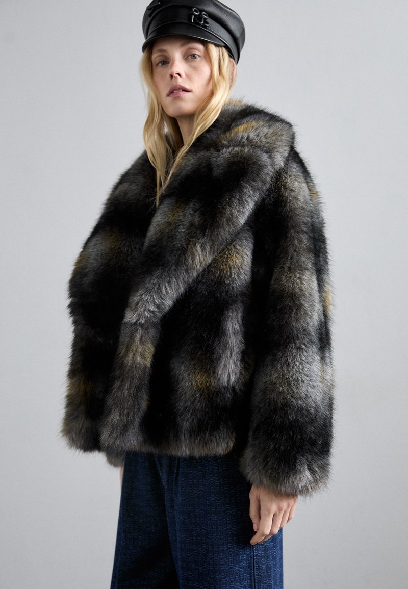 Blondynka w ciemnym, wielokolorowym futrze, czapce skórzanej w czarnym kolorze oraz wzorzystych, ciemnonawigacyjnych spodniach, stojąca na jednolitym szarym tle.