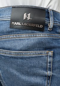 Μπλε τζιν παντελόνι με φθαρμένη υφή, που διαθέτει μαύρο δερμάτινο ταμπελάκι στη μέση με ανάγλυφο το "KARL LAGERFELD" και λεπτομέρειες από ασημένια μεταλλικά στοιχεία.