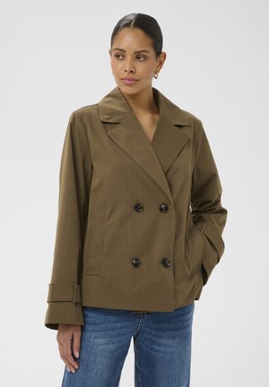 Femme portant une veste olive à double boutonnage avec de larges revers et un jean bleu, une main dans la poche, sur un fond uni.