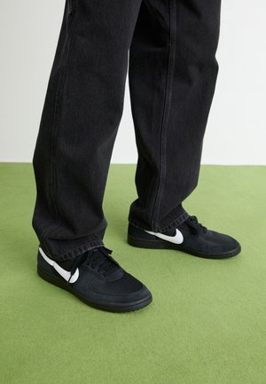 Osoba w czarnych dżinsach i czarnych butach Nike z białym znakiem Swoosh stoi na zielonej podłodze przy białej ścianie.