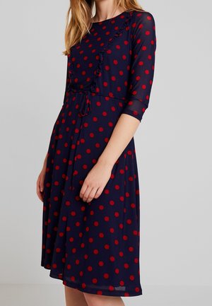 Frau trägt ein marineblaues Kleid mit roten Polka-Dots, dreiviertel Ärmeln, einem schrägen Rüschendetail und einem Taillengürtel, steht vor einem schlichten Hintergrund.