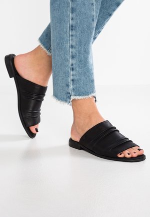 Füße in hellblauen Jeans mit ausgefranstem Saum, die schwarze offene Leder-Slipper tragen, auf weißem Boden.