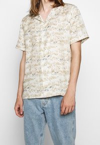 Camicia beige a maniche corte con un motivo floreale, caratterizzata da dettagli in azzurro chiaro e blu scuro, chiusura con bottoni e vestibilità rilassata.