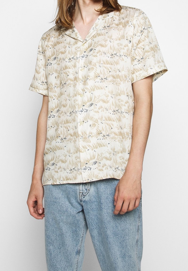 Camicia beige a maniche corte con un motivo floreale, caratterizzata da dettagli in azzurro chiaro e blu scuro, chiusura con bottoni e vestibilità rilassata.
