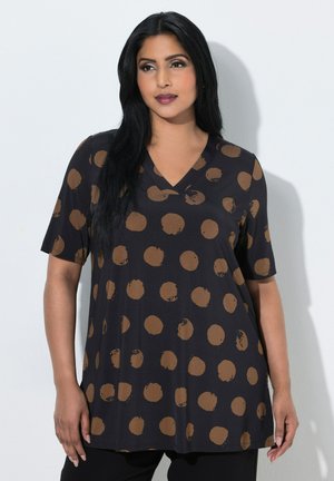 Vrouw met lang zwart haar, die een zwart V-hals shirt met grote bruine stippen draagt, staand tegen een witte achtergrond.