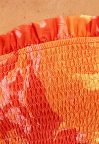 Tissu smocké à motif floral orange vif avec un bord à volants ; présente des coutures texturées et froncées ainsi qu'une apparence douce et légère.