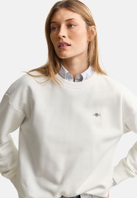 Jeune femme aux longs cheveux châtain clair portant un sweat blanc par-dessus une chemise bleu clair boutonnée, regardant sur le côté avec les mains sur les hanches.