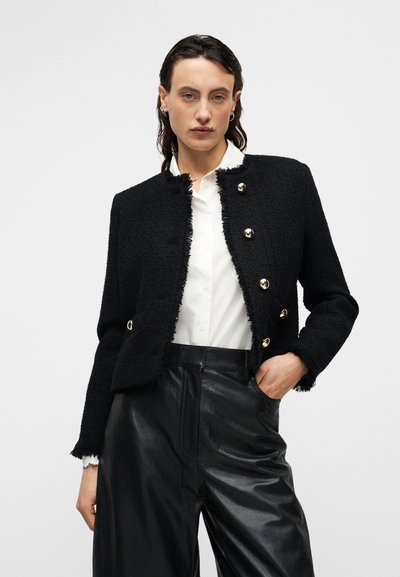Μαύρο κοντό σακάκι από υφασμάτινη υφή με fringing στις άκρες, με κλεισίματα από χρυσά κουμπιά. Συνδυασμένο με μαύρο δερμάτινο παντελόνι ψηλόμεσο και λευκό πουκάμισο.
