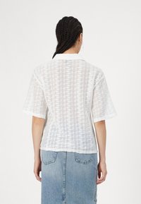 Camicia bianca a maniche corte con un motivo a maglia aperta e testurizzata, caratterizzata da un colletto classico e una vestibilità leggermente ampia. Abbinata a una gonna in denim.