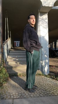 Jaunas vyras, vilkintis juodą džemperį su gobtuvu ir žalias sportines kelnes su baltomis juostelėmis, stovi su rankomis kišenėse prie laiptų po betono konstrukcija.