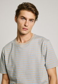 Raižyta marškinėliai smėlio ir šviesiai mėlynos spalvos, pagaminti iš medvilninio audinio. Yra apvalus kaklas ir siuvinėtas logotipas ant kairės krūtinės.