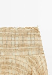 Beiger Stoff mit einem strukturierten Karomuster. Das Material hat eine leicht grobe Haptik, mit hellen und dunklen Linien, die ein Rautendesign bilden.