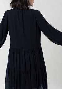 Mustaas tikkidress, millel on pikad läbipaistvad varrukad ja seljaosast võtmeaugu detail. Valmistatud kerge ja pehme tekstuuriga kangast.