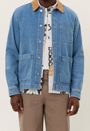 Homme portant une veste en jean bleu clair avec un col beige par-dessus une chemise à motifs et un pantalon marron, debout devant un fond uni.