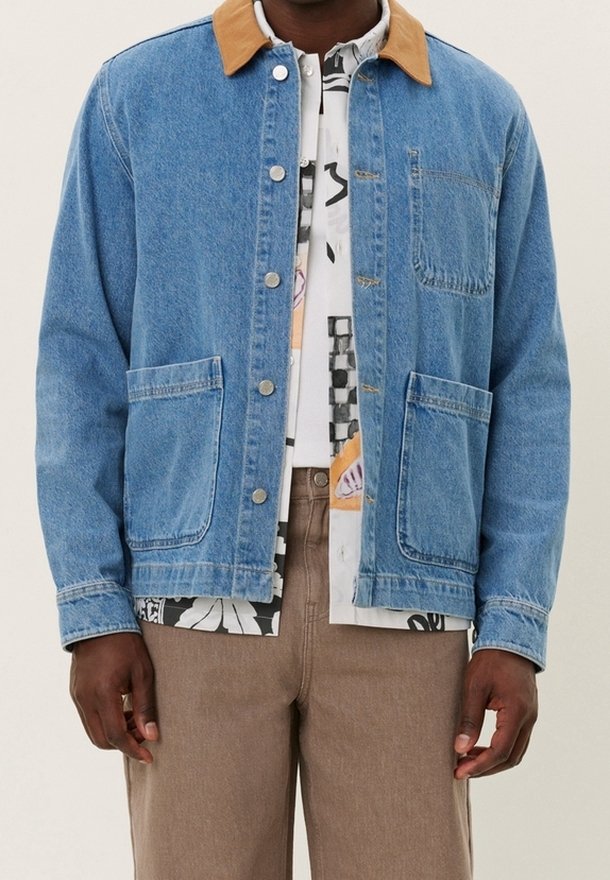 Homme portant une veste en jean bleu clair avec un col beige par-dessus une chemise à motifs et un pantalon marron, debout devant un fond uni.