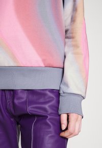 Sudadera multicolor con un patrón degradado en tonos rosa, púrpura y gris, con un puño acanalado gris y una textura suave. Combinada con pantalones de cuero púrpura.