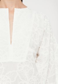 Fehér vászonblúz V-kivágással, finom virágmintás hímzéssel és hosszú, enyhén puffos ujjakkal. Textúrált részletezés az egész anyagon.