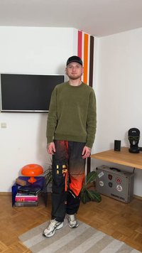 Camisola verde-oliva, calças com padrão preto e laranja, sapatos cinzentos. Texturas suaves, corte descontraído. Acessórios incluem um boné preto.