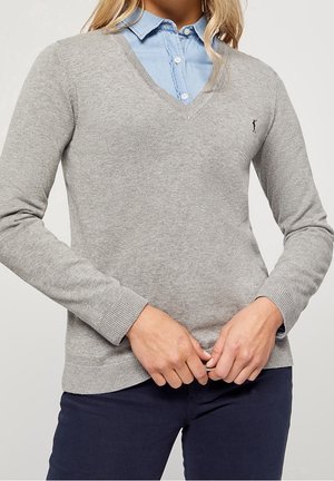 Femme portant une chemise bleu clair boutonnée sous un pull gris col en V avec un pantalon bleu marine, mains jointes à la taille.