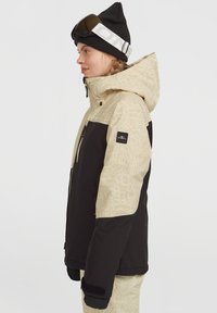 Skijack met een mix van beige met patroon en zwarte panelen, voorzien van een ritssluiting aan de voorkant, een verstelbare capuchon en logo-accenten. Warme, gestructureerde stof.