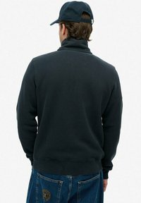 Sudadera de color azul marino con cuello alto y puños de canalé, hecha de una tela suave. Llevada con pantalones de mezclilla azul y una gorra a juego.