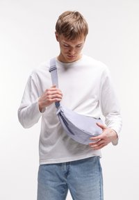 Vaaleanharmaa olkalaukku, joka on valmistettu kankaasta ja jossa on pyöreä muoto, säädettävä olkahihna ja hillitty logomerkintä. Kantaa vartalon yli.