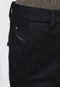 Svarta denimbyxor med en slät textur, slim fit, femficksdesign och en distinkt blå metallknapp fram.