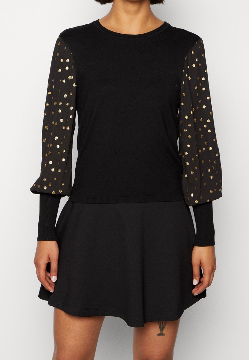 Haut noir à manches longues avec des manches bouffantes transparentes ornées de pois dorés, associé à une jupe noire, porté par une personne debout.