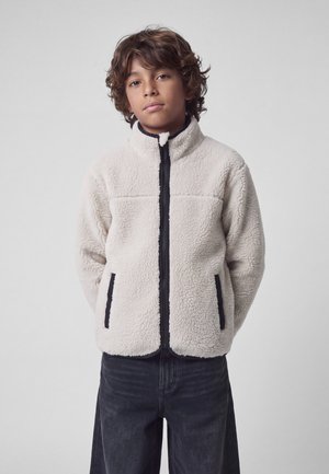 Garçon aux cheveux bouclés portant une veste polaire blanche avec une fermeture éclair noire et des poches, debout devant un fond gris uni.