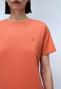 Oranges T-Shirt aus Baumwolle mit kurzen Ärmeln, das ein kleines gesticktes Logo auf der Brust präsentiert. Rundhalsausschnitt und glatte Textur.