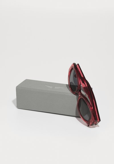 Lunettes de soleil rouges avec des montures angulaires surdimensionnées reposant sur une boîte gris clair. Les verres sont sombres et les montures présentent une finition brillante.