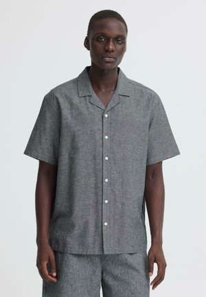 Homme portant une chemise grise à manches courtes boutonnée avec un short gris assorti sur un fond clair uni.