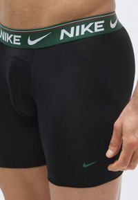 Boxeri negri cu logo-ul Nike verde și alb pe banda elastică a taliei. Țesătură netedă cu design ajustat pe corp.