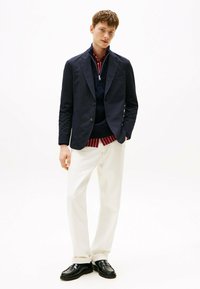 Blazer blu navy sopra un maglione scuro con colletto, abbinato a pantaloni bianchi e scarpe nere. L'outfit presenta linee pulite e una vestibilità su misura.