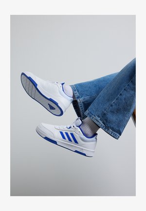 Zapatillas deportivas blancas con acentos azules, que cuentan con laterales perforados, una marca de tres franjas y una suela de goma texturizada. Combinadas con jeans azul claro.