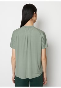 Top de manga corta en verde claro, con cuello redondo y detalles fruncidos en la parte posterior. Tejido suave con una textura lisa.