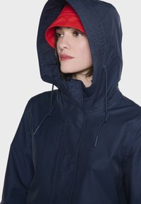 Mørkeblå vandtæt jakke med snor i hætten, der har en rød inderside. Glat tekstur og minimalistisk design uden synligt hardware.
