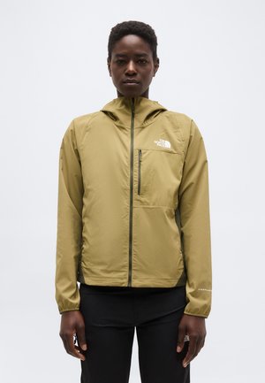 Olíva zöld The North Face kapucnis cipzáras dzsekit viselő személy, világos, egyszínű háttér előtt áll.